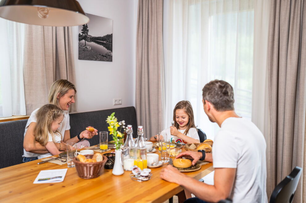 Familie beim Frühstück, lächelnd und genießend, mit frischen Brötchen und Säften.