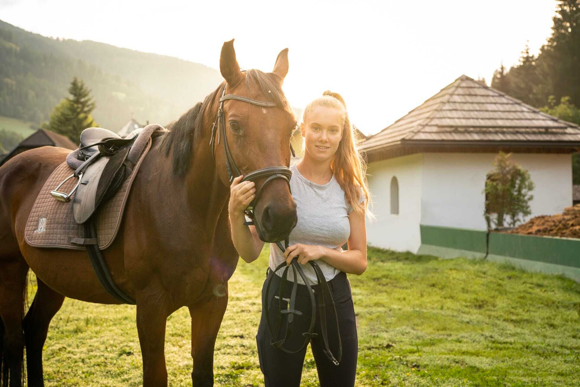Woman with horse at sunset on a green meadow in front of a chalet.