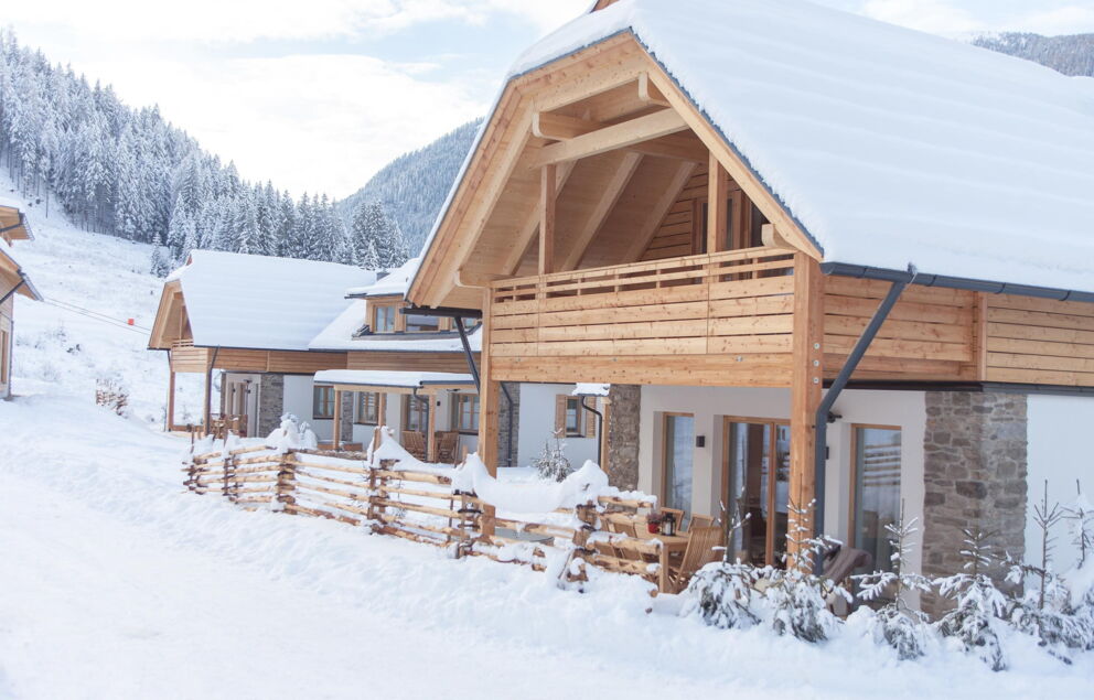 Verschneite Chalets mit Holzdetails vor majestätischen Bergen im Winterpanorama.