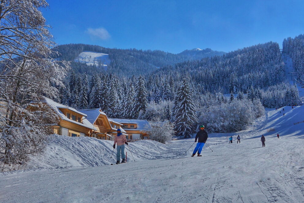 Skifahrer in verschneiter Landschaft nahe gemütlicher Chalets mit bewaldeten Hügeln im Hintergrund.