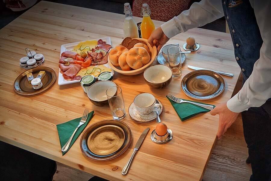 Frühstückstisch mit frischen Brötchen, Aufschnitt, Getränken und gedecktem Geschirr.