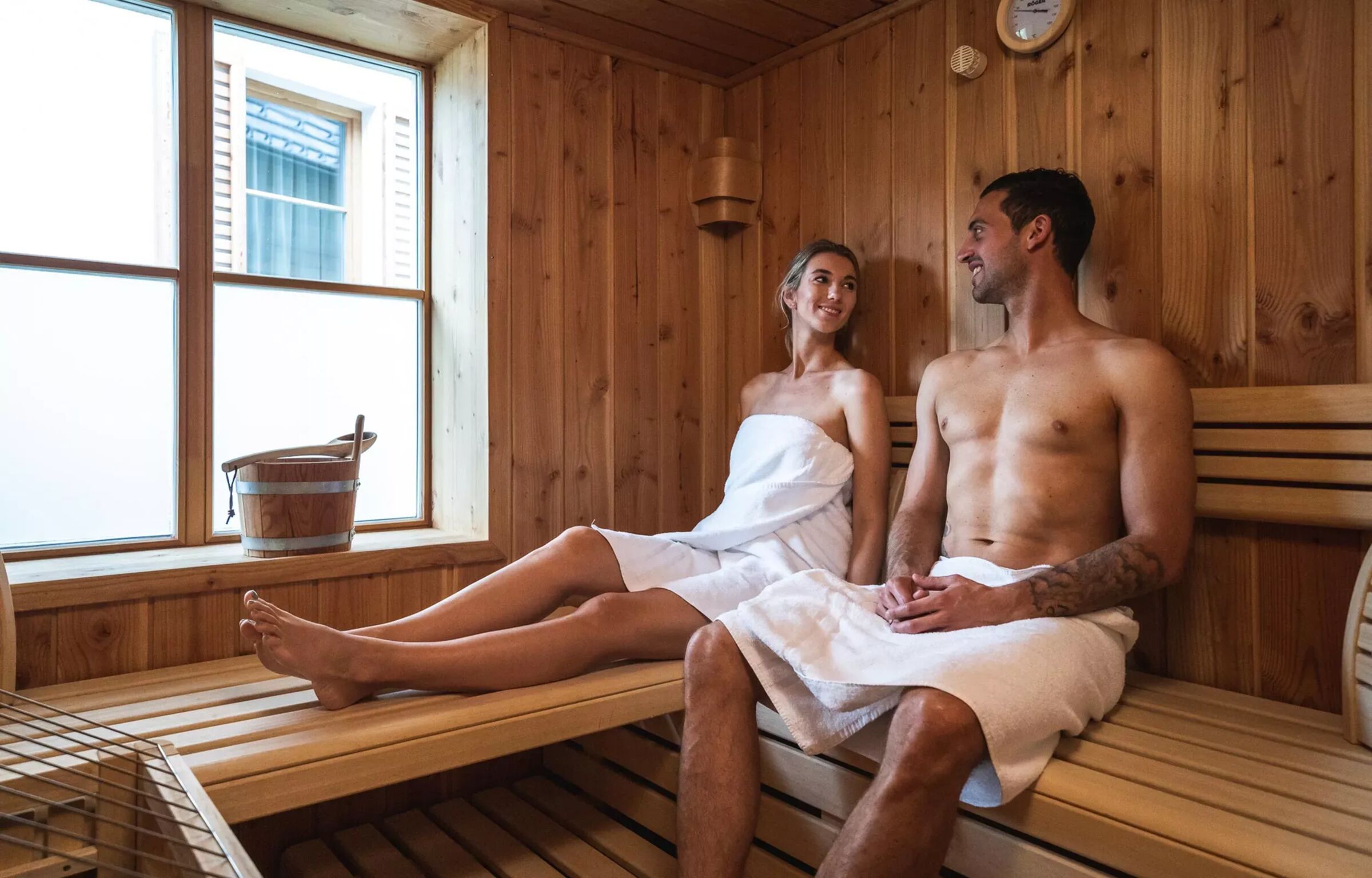 Zwei Personen entspannen in einer hölzernen Sauna mit Fensterblick.