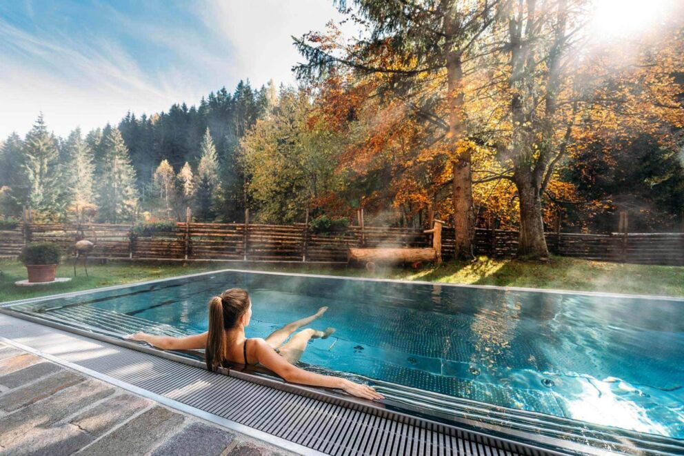 Woman relaxing in the outdoor pool, surrounded by an autumnal forest landscape.
