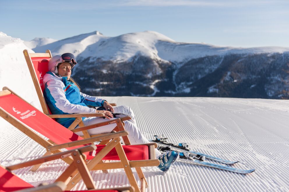 Frau entspannt auf Liegestuhl im Schnee mit Bergblick und Skiern.