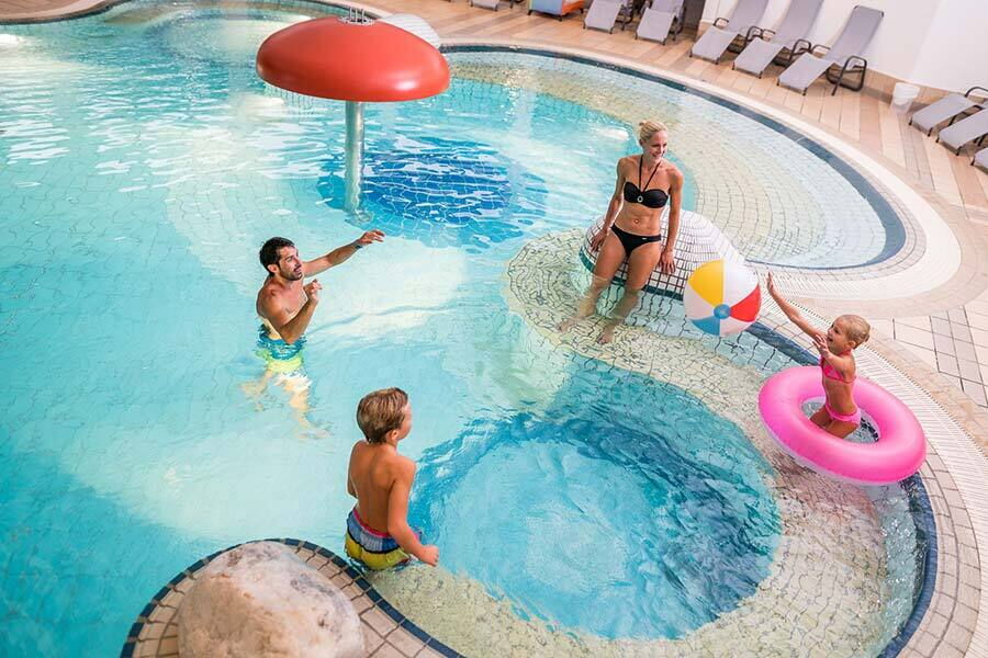 Family with children playing in the indoor pool with colorful swimming rings and water mushroom.
