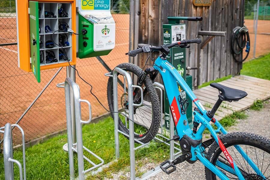 E-bike parked at charging station in front of a tennis court and wooden building.