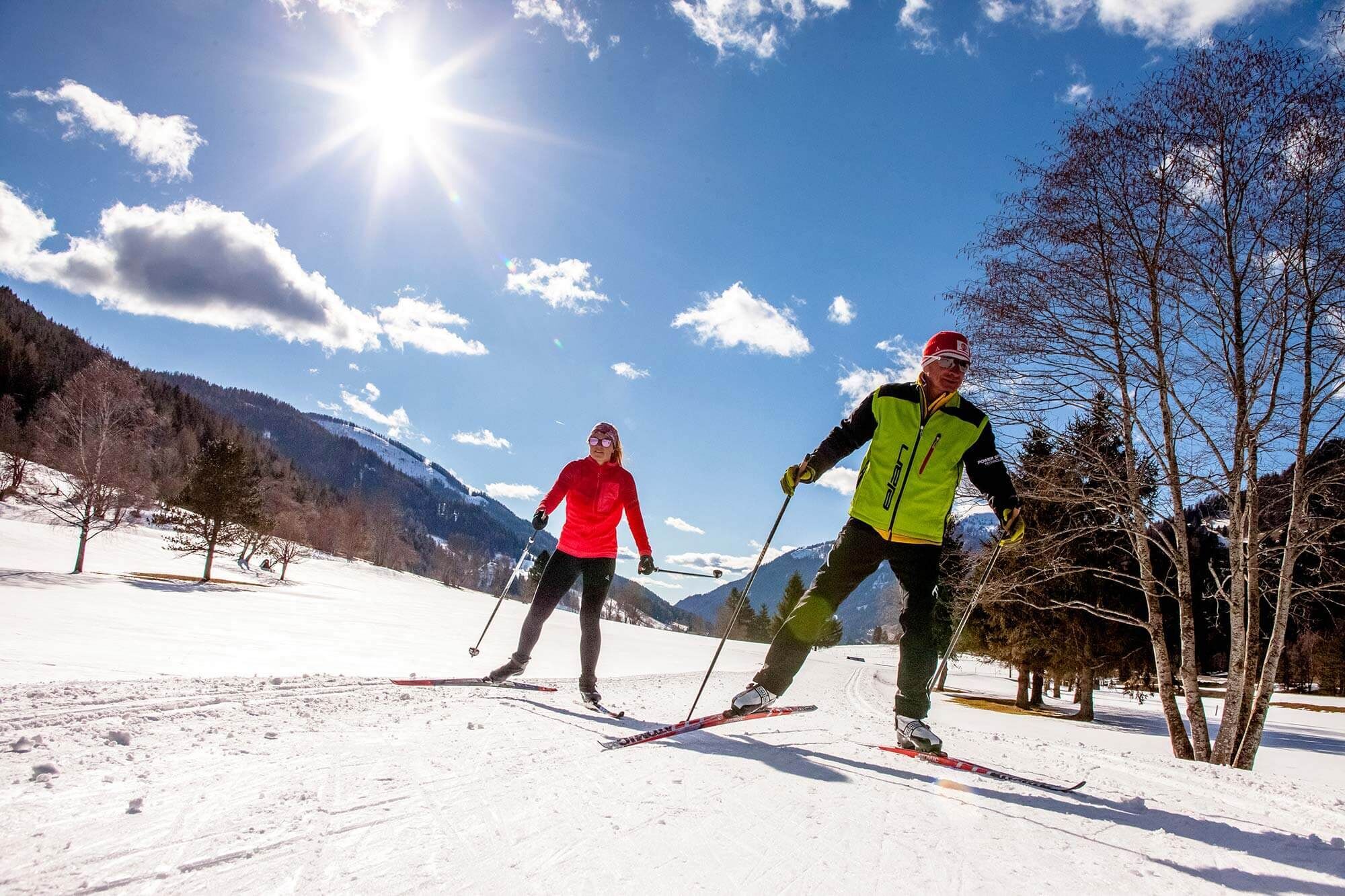 Two cross-country skiers enjoy the snow-covered landscape under bright sunshine and blue skies.
