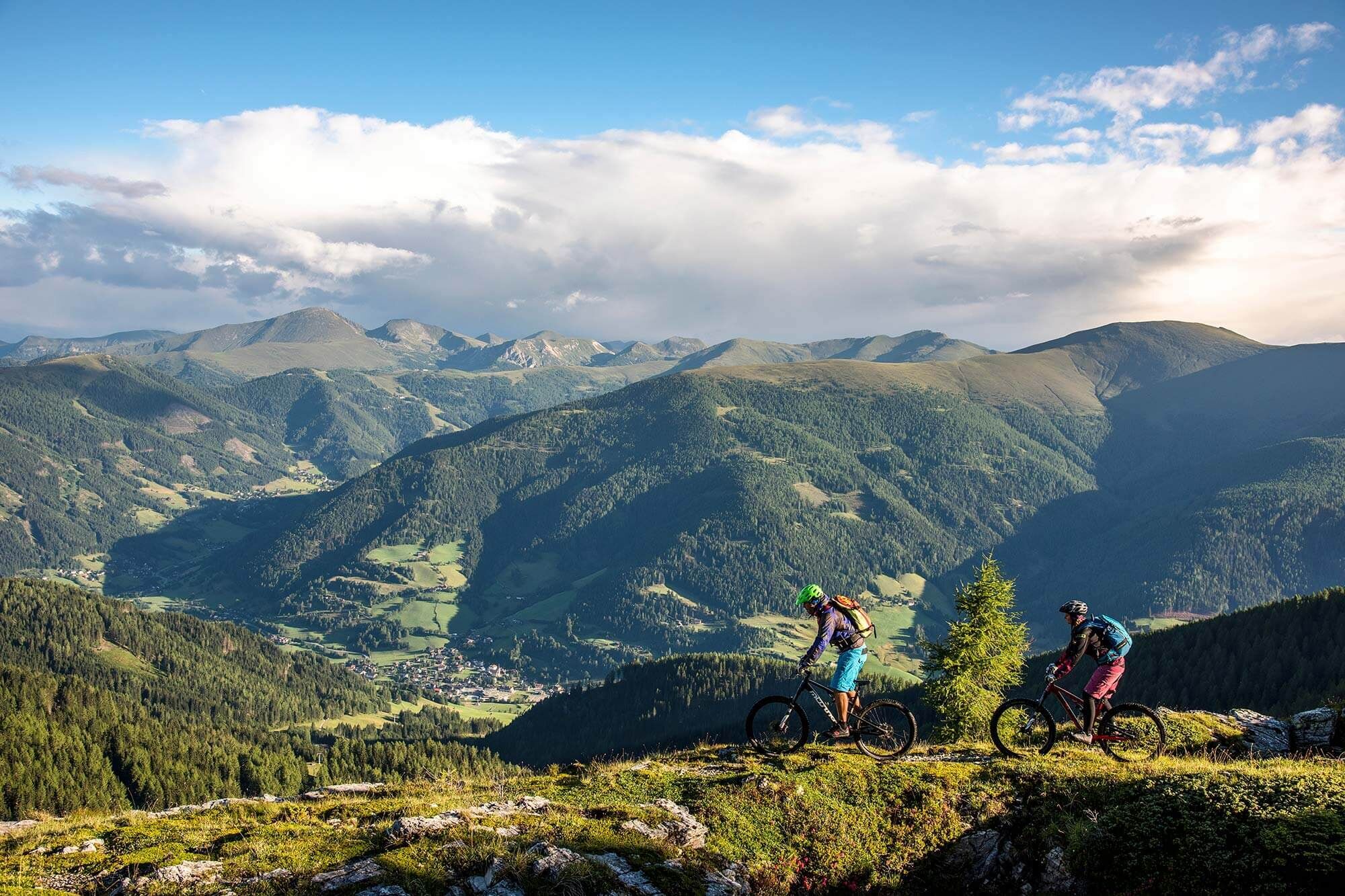 Two mountain bikers ride along a mountain trail with breathtaking views of the Nockberge mountains.