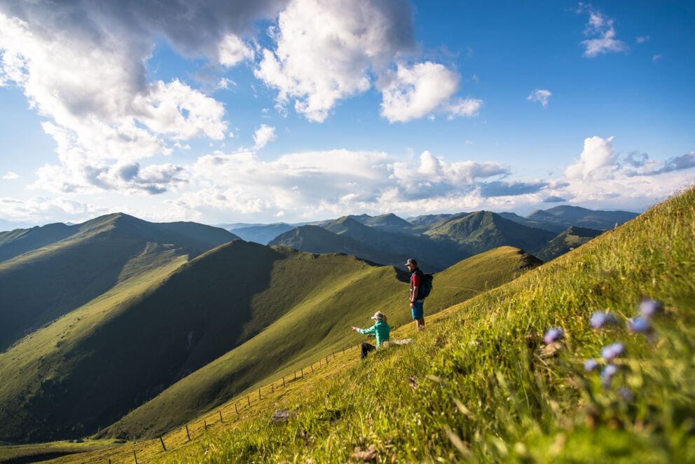 Zwei Personen wandern auf einem sonnigen Bergpfad mit weitem Blick auf die Nockberge.