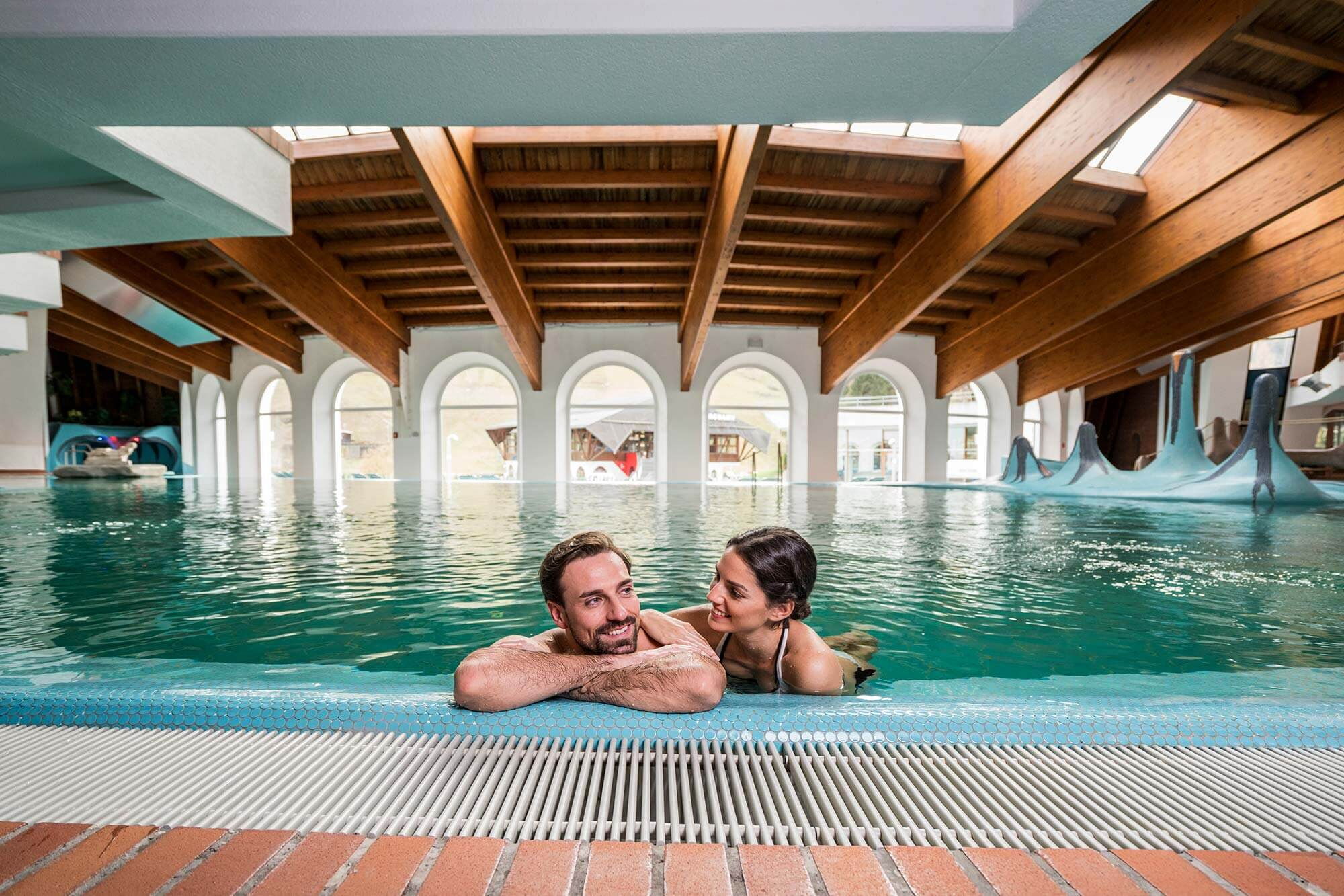 La coppia si rilassa nella piscina termale sotto soffitti in legno con grandi finestre e luce naturale.