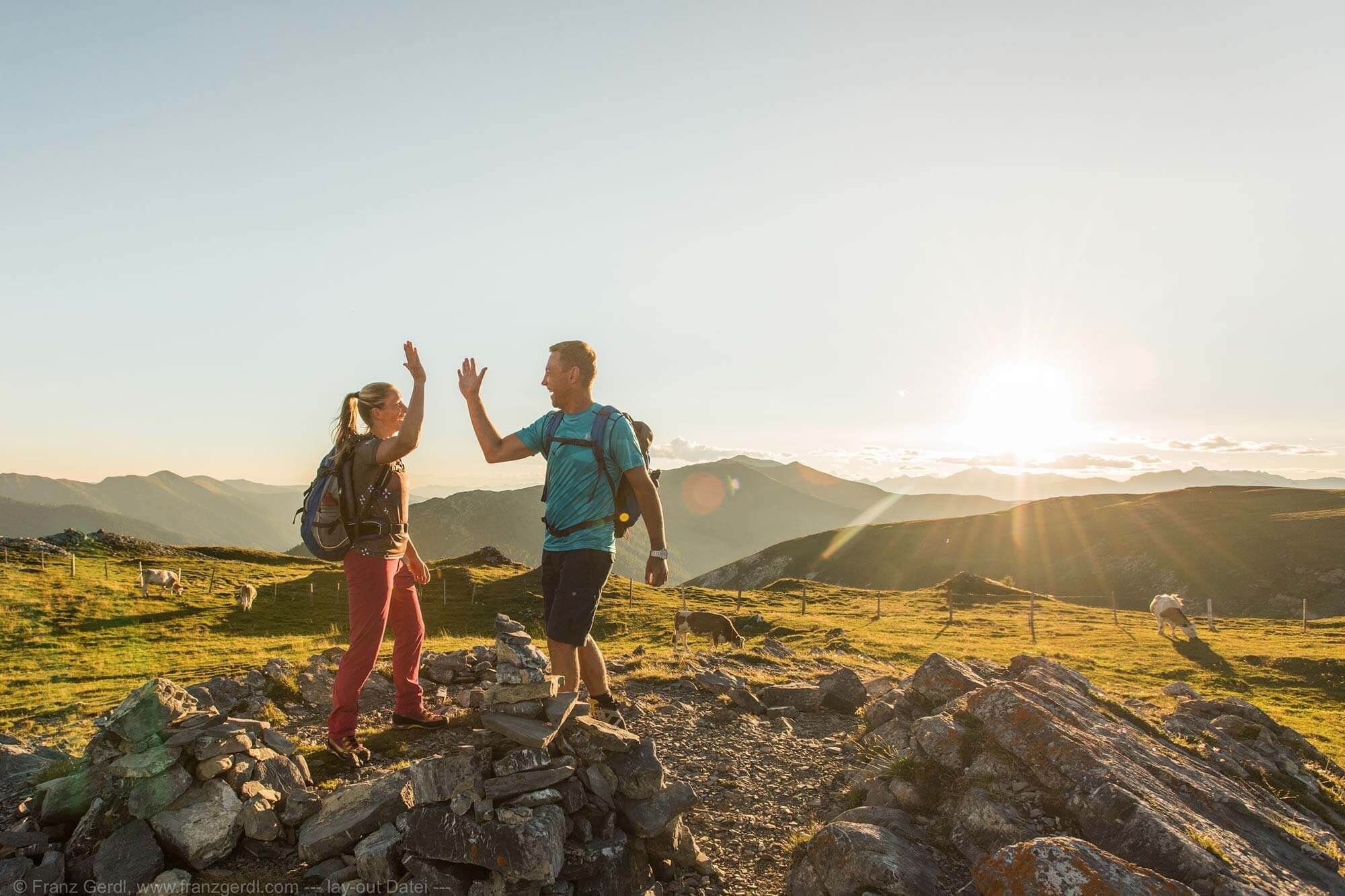 Two hikers give each other a high-five, the sun rises over the Nockberge mountains.