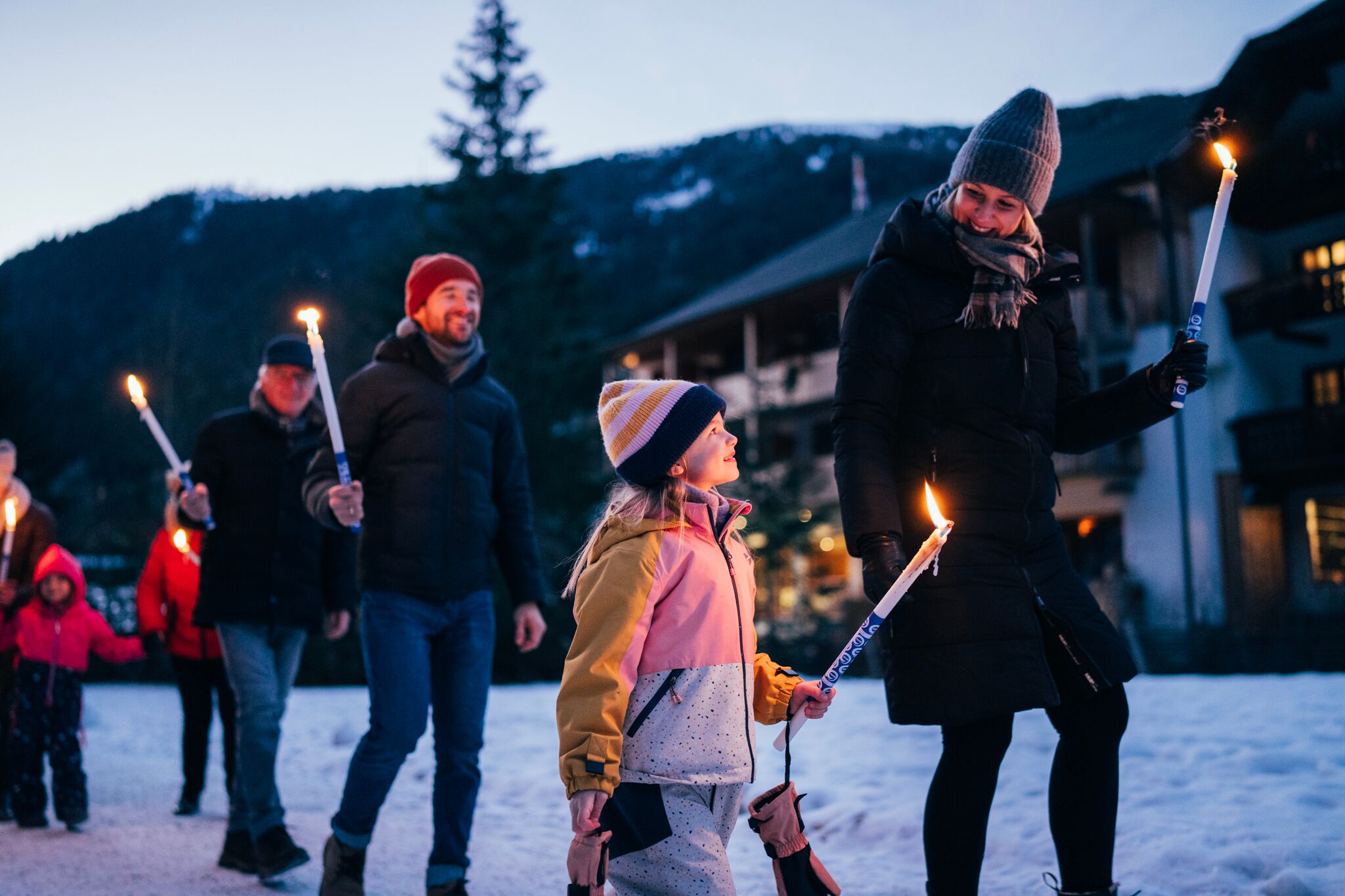 Persone che camminano nella neve con le torce, sorridenti e in abiti invernali.