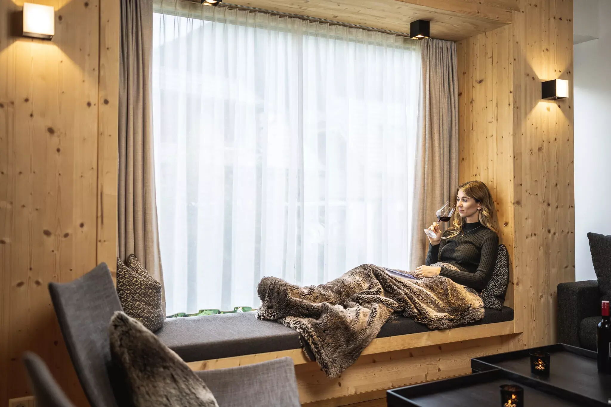 Frau entspannt mit Glas Wein auf gemütlicher Fensterbank mit Decke.