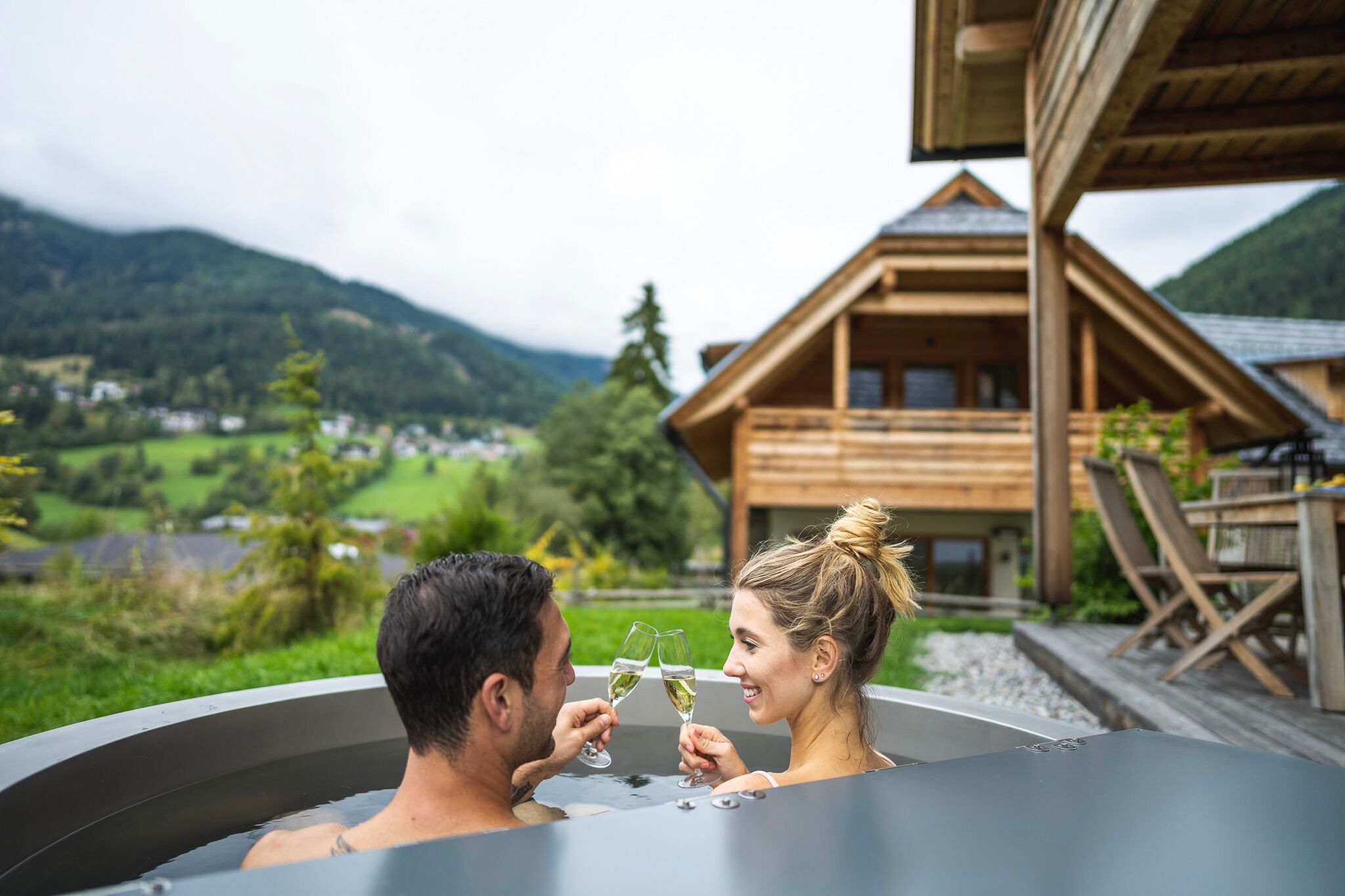 Coppia che si rilassa nella vasca idromassaggio con champagne, circondata da paesaggi alpini e chalet.
