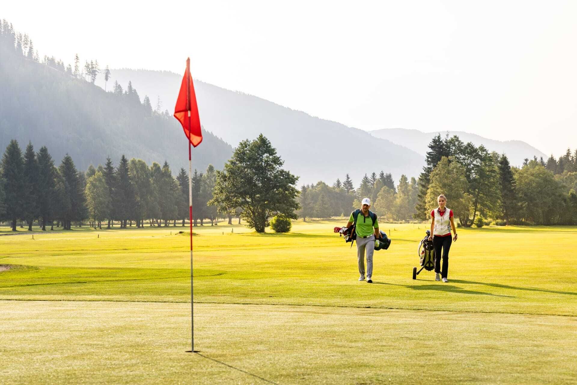 Zwei Golfspieler auf grünem Platz, umgeben von Bergen und Bäumen.