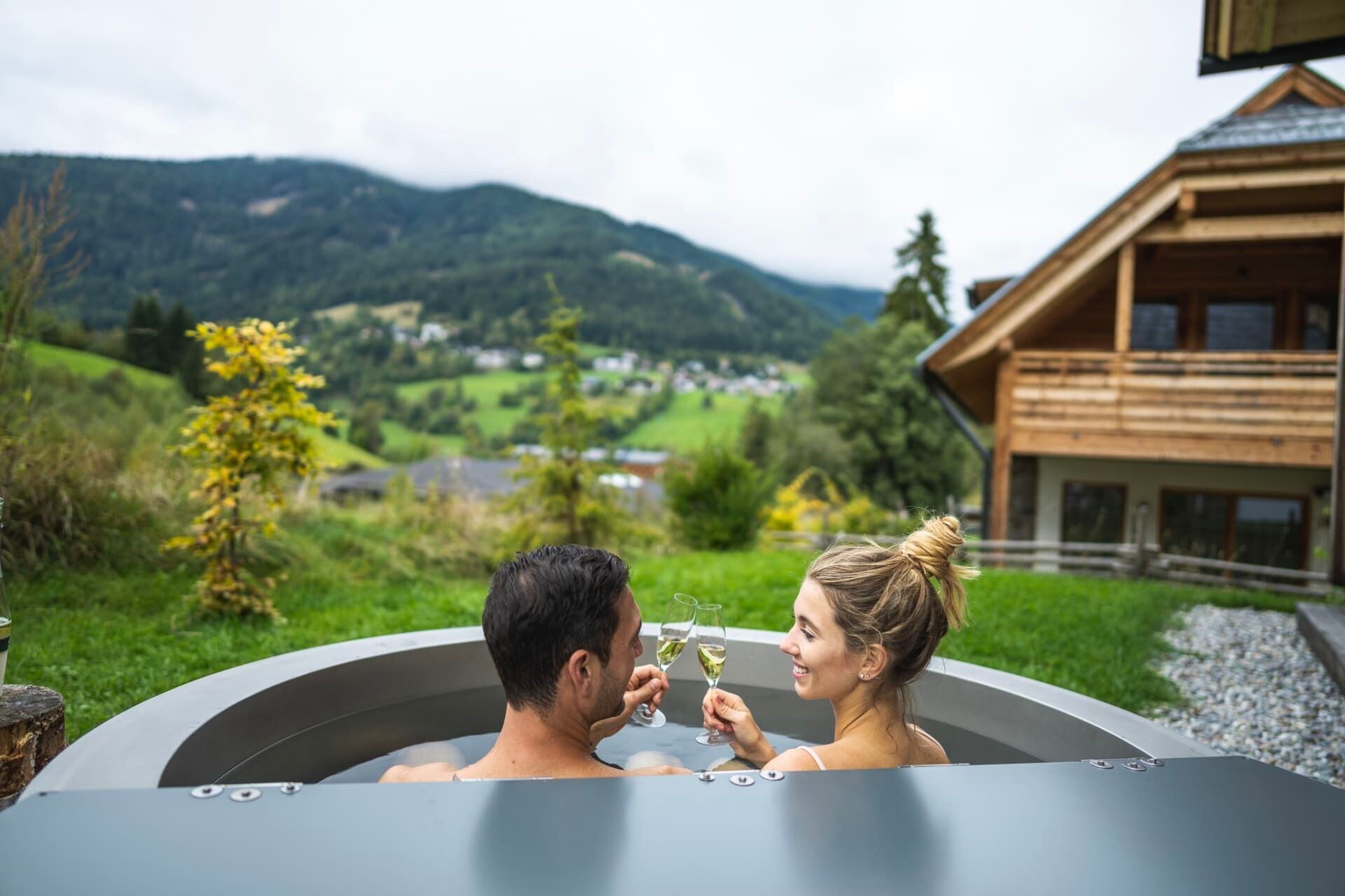 Coppia che si rilassa nella vasca idromassaggio con lo champagne, con il pittoresco paesaggio montano e gli chalet sullo sfondo.
