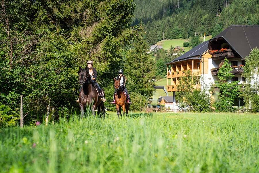 Zwei Reiter auf Pferden vor einem Chalet in grüner Landschaft