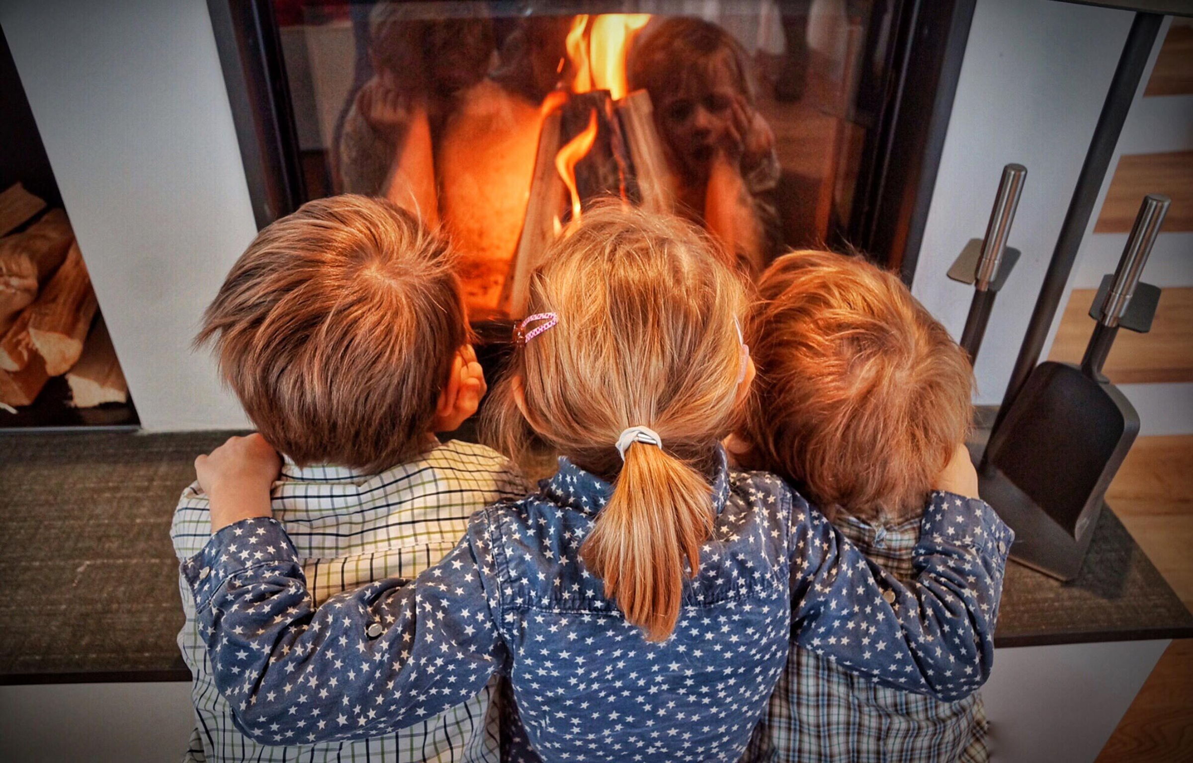 Drei Kinder sitzen eng umarmt vor einem lodernden Kaminfeuer.