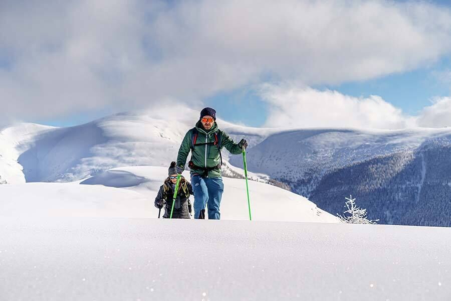 Two people hike through a snow-covered landscape in front of majestic mountain peaks and a blue sky.