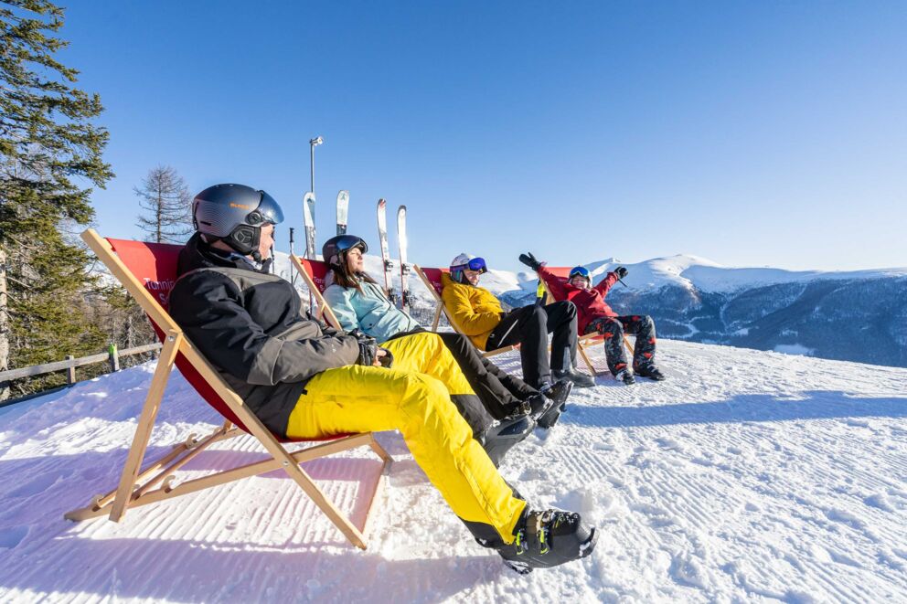 Menschen entspannen in Liegestühlen im Schnee mit Bergblick und strahlend blauem Himmel.