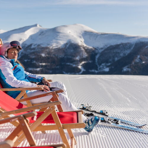 Frau entspannt auf Liegestuhl im Schnee mit Bergblick und Skiern.