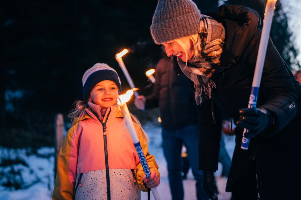 Adulti e bambini durante una fiaccolata serale nella neve, sorridenti e felici.