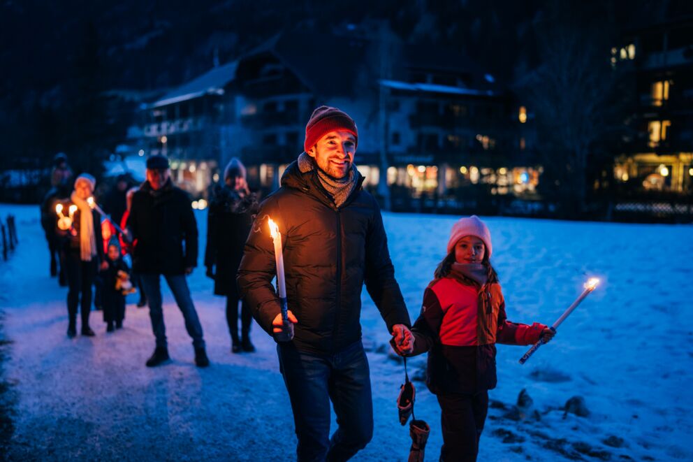 Menschen in Winterkleidung bei einer Fackelwanderung im Schnee in den Abendstunden.