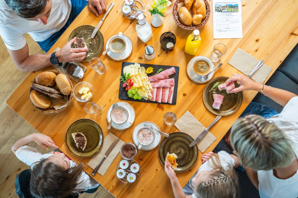 Familie beim gemeinsamen Frühstück mit Brötchen, Aufschnitt und Getränken.