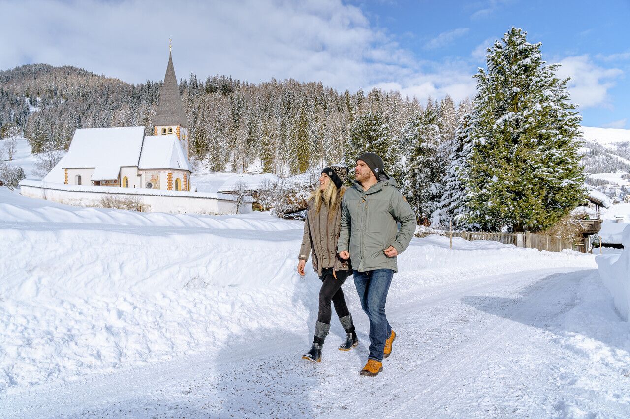 Couple walking through a snowy landscape with a church in the background under a clear sky.