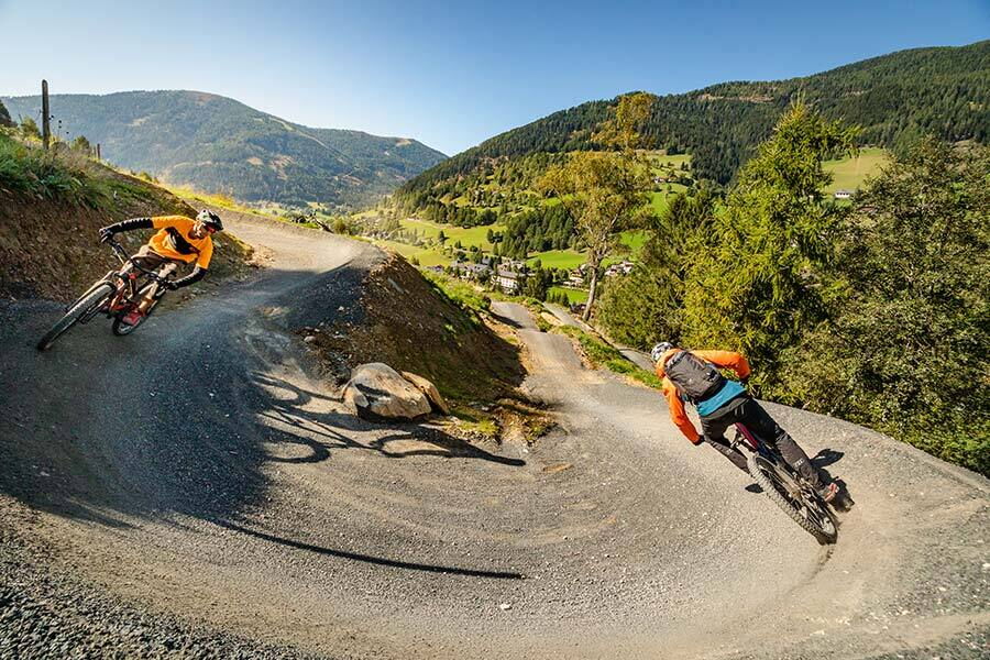 Zwei Mountainbiker auf einem Flowtrail mit malerischer Bergkulisse im Hintergrund.
