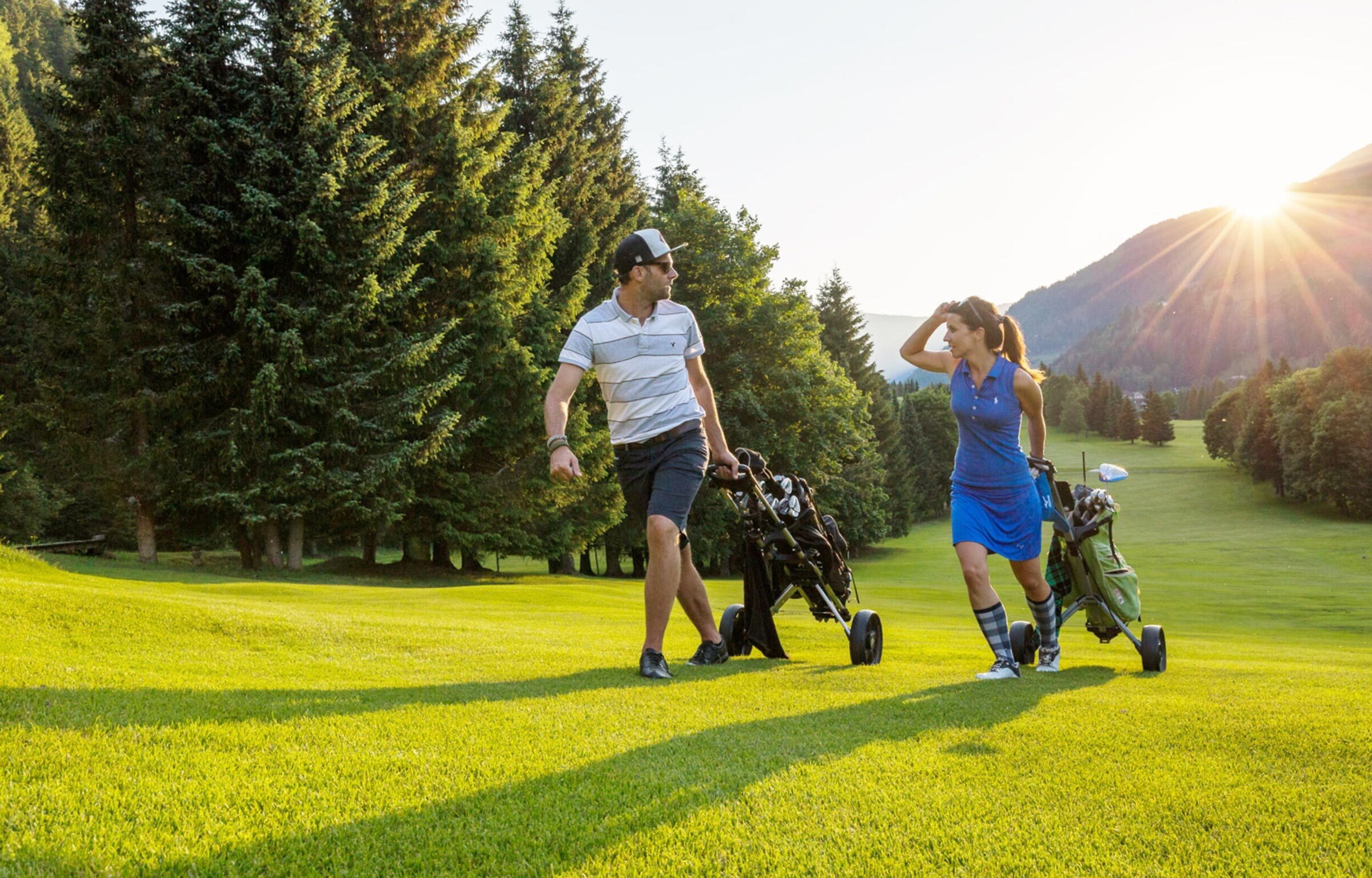 Due persone che giocano a golf su un campo verde, circondato da alberi e montagne.