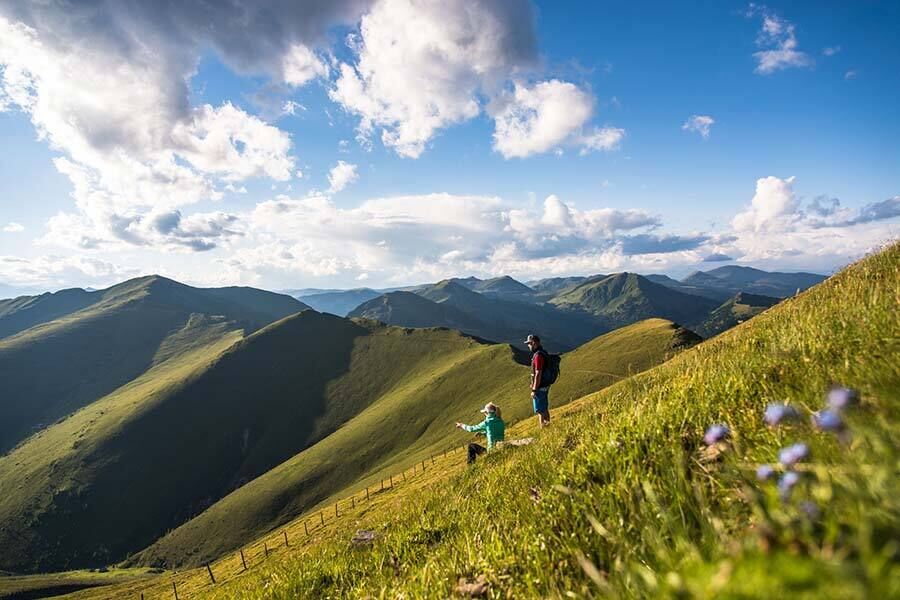 Two people hike over green hills with sweeping views of the Nockberge mountains.