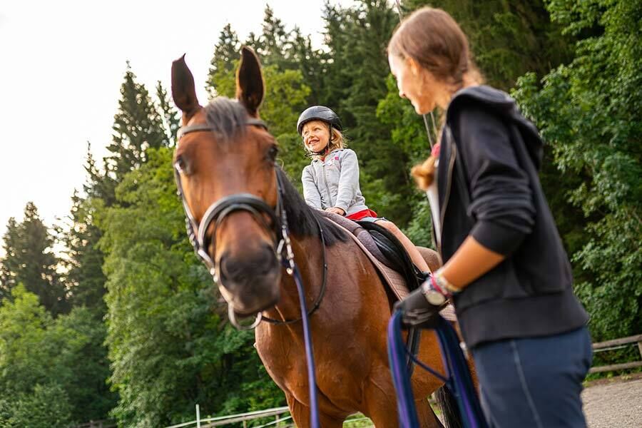Child smiling on horse, looked after by a woman in the forest.