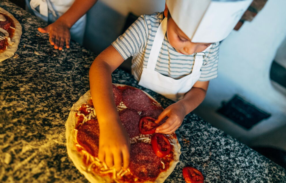 Child creates pizza with fresh ingredients on a dark worktop.