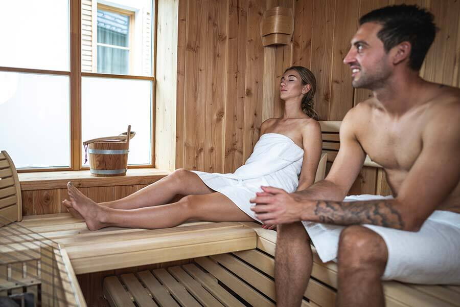 Zwei Personen entspannen in einer Sauna mit Holzbänken und Fensterblick.