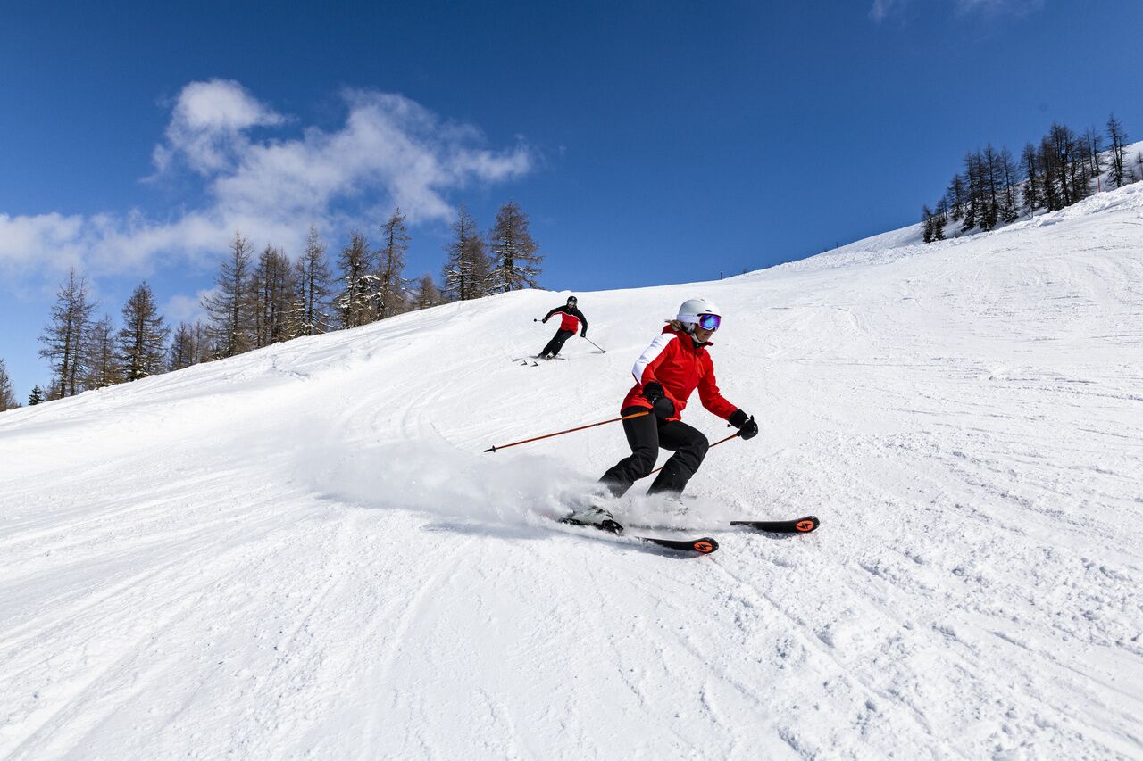 Zwei Skifahrer auf schneebedeckter Piste unter klarem, blauem Himmel.