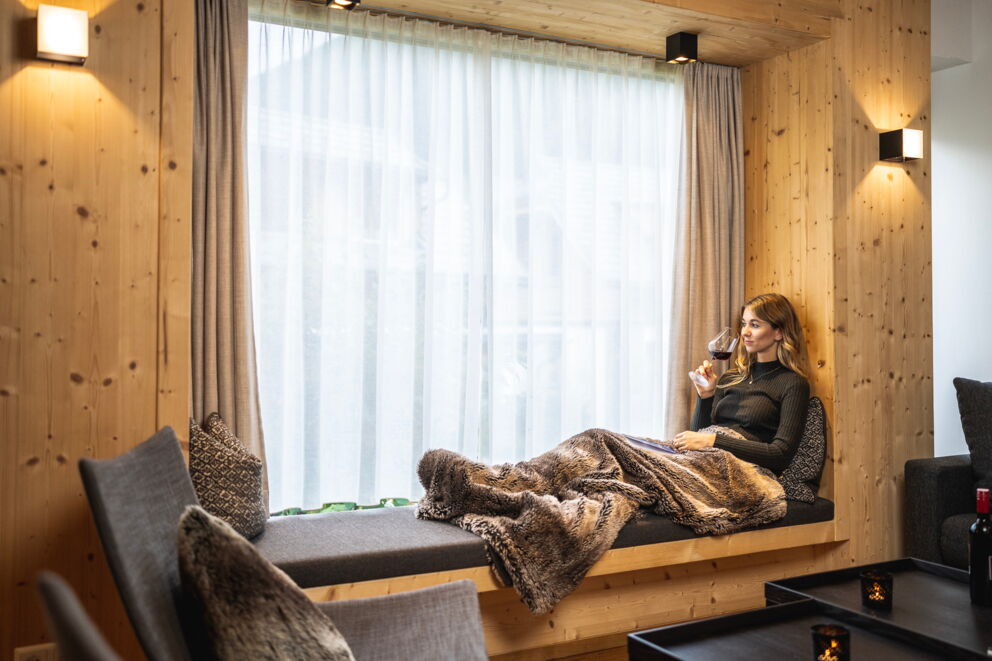 Frau genießt Wein auf gemütlicher Sitzbank im Chalet mit weichem Fell und Kerzenlicht.