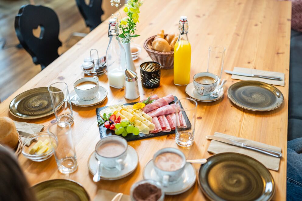 Frühstückstisch mit Aufschnitt, Saft, Kaffee und frischen Brötchen auf Holzplatte.