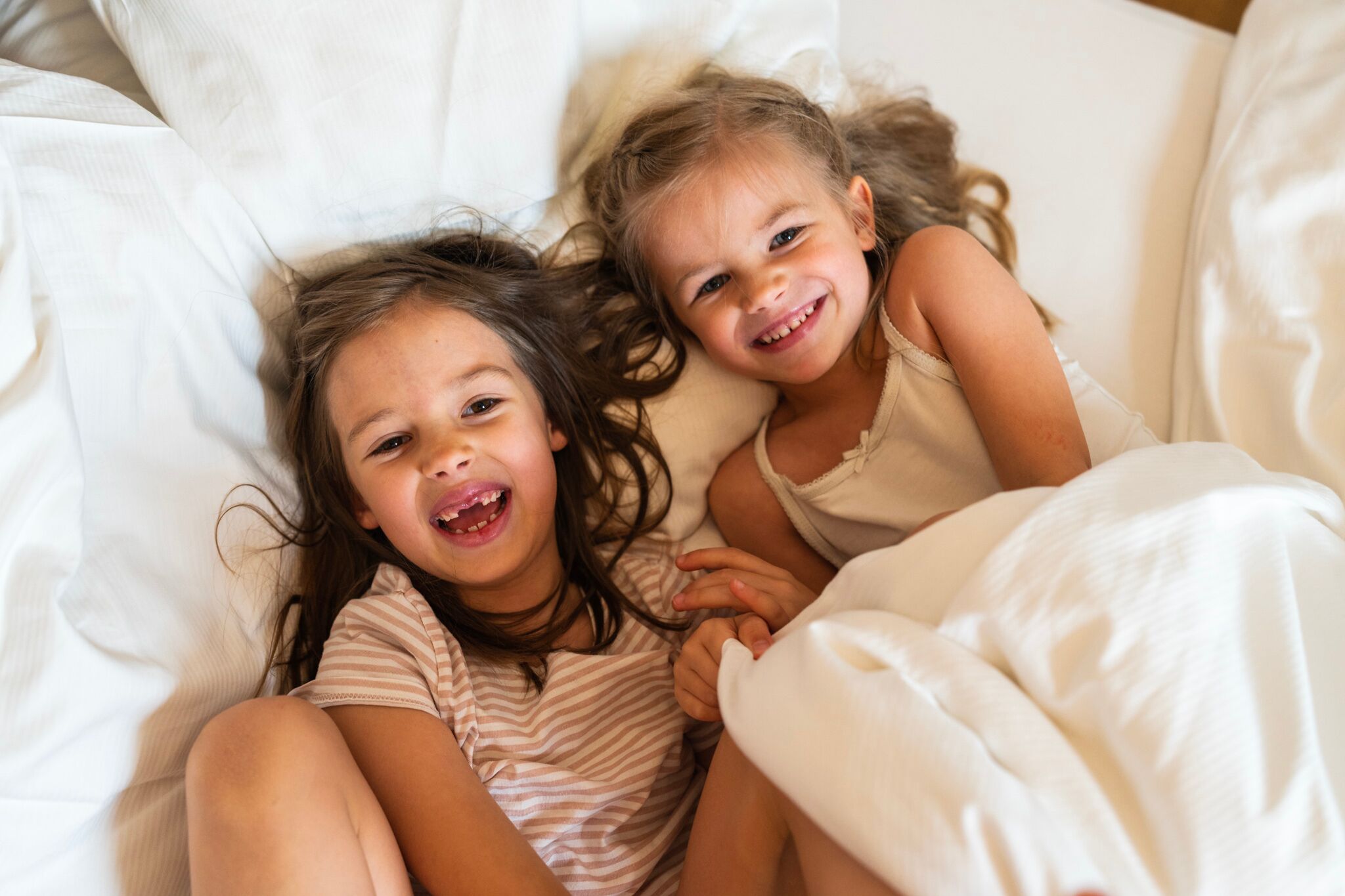 Due bambini che ridono, rannicchiati in accoglienti piumoni bianchi.