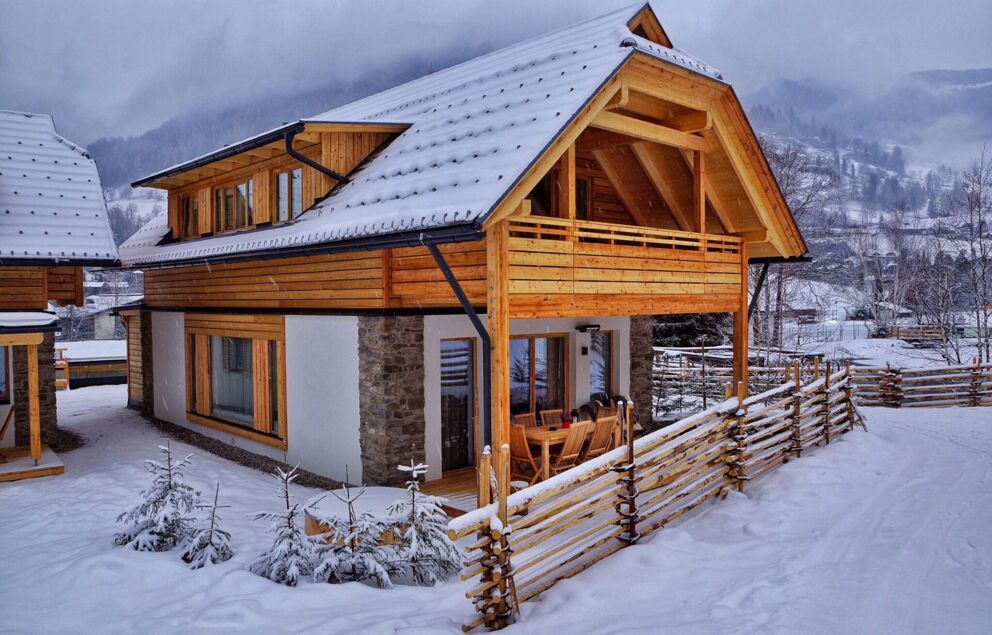 Schneebedecktes Holzchalet mit Balkon und eingezäuntem Garten in malerischer Winterlandschaft.