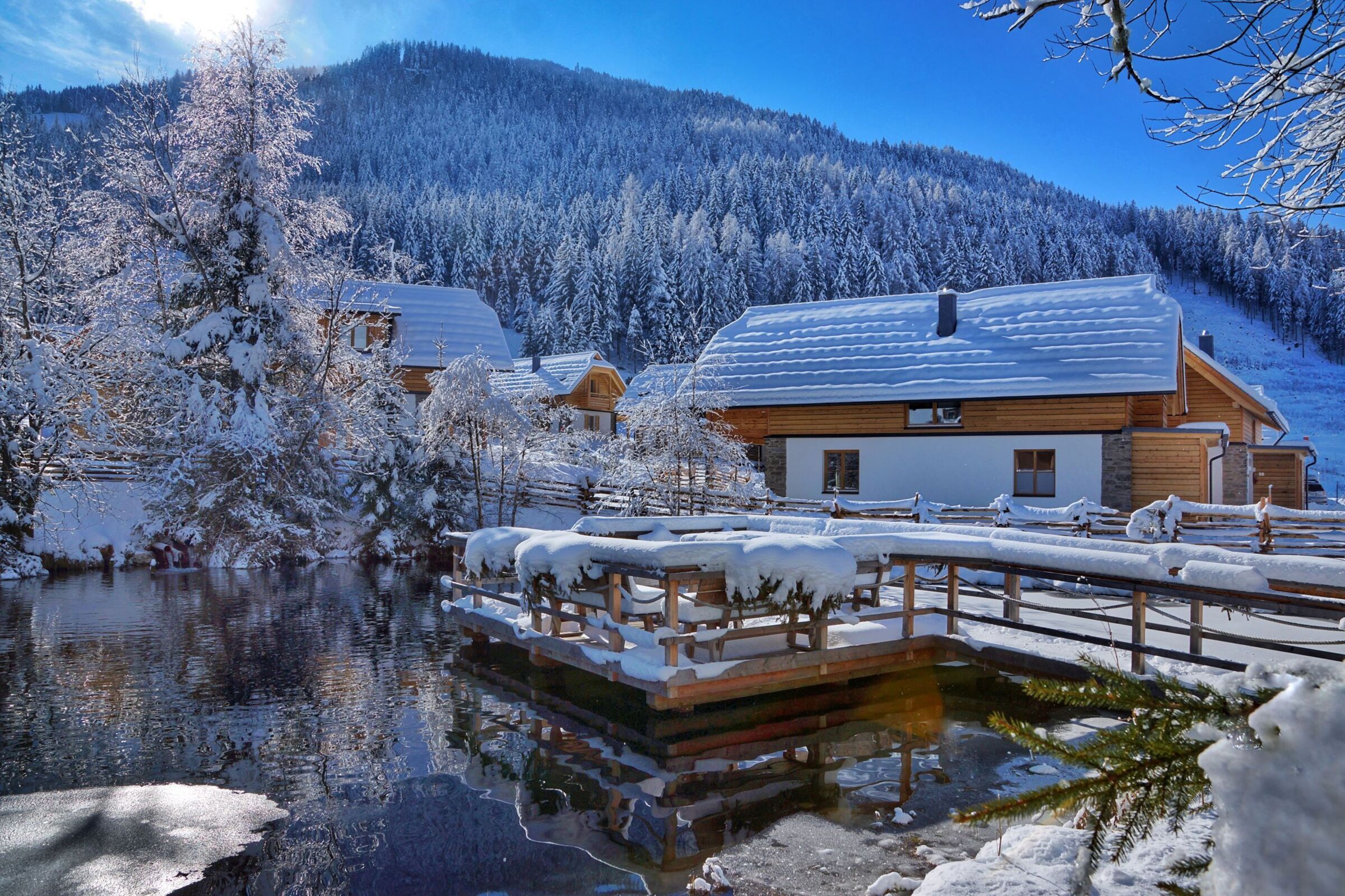 Chalet innevati accanto a un laghetto scintillante, circondati da alberi e montagne innevate.