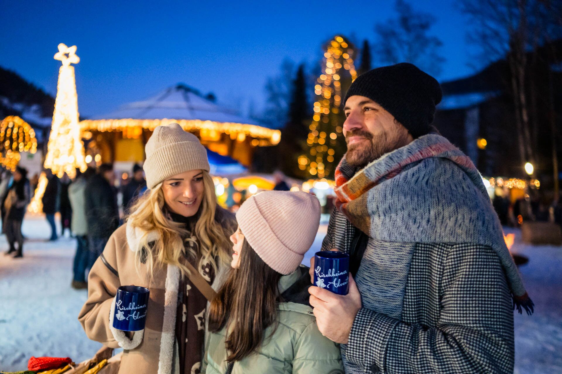 Familie genießt winterlichen Markt mit Lichterketten und heißem Getränk in der Hand.
