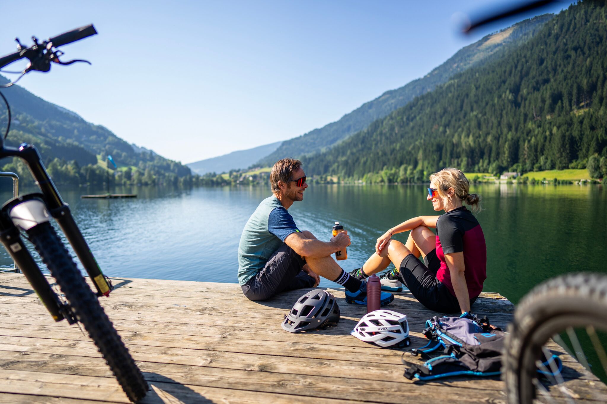 Two cyclists relax on a jetty on the sunny lakeshore with a view of the mountains.