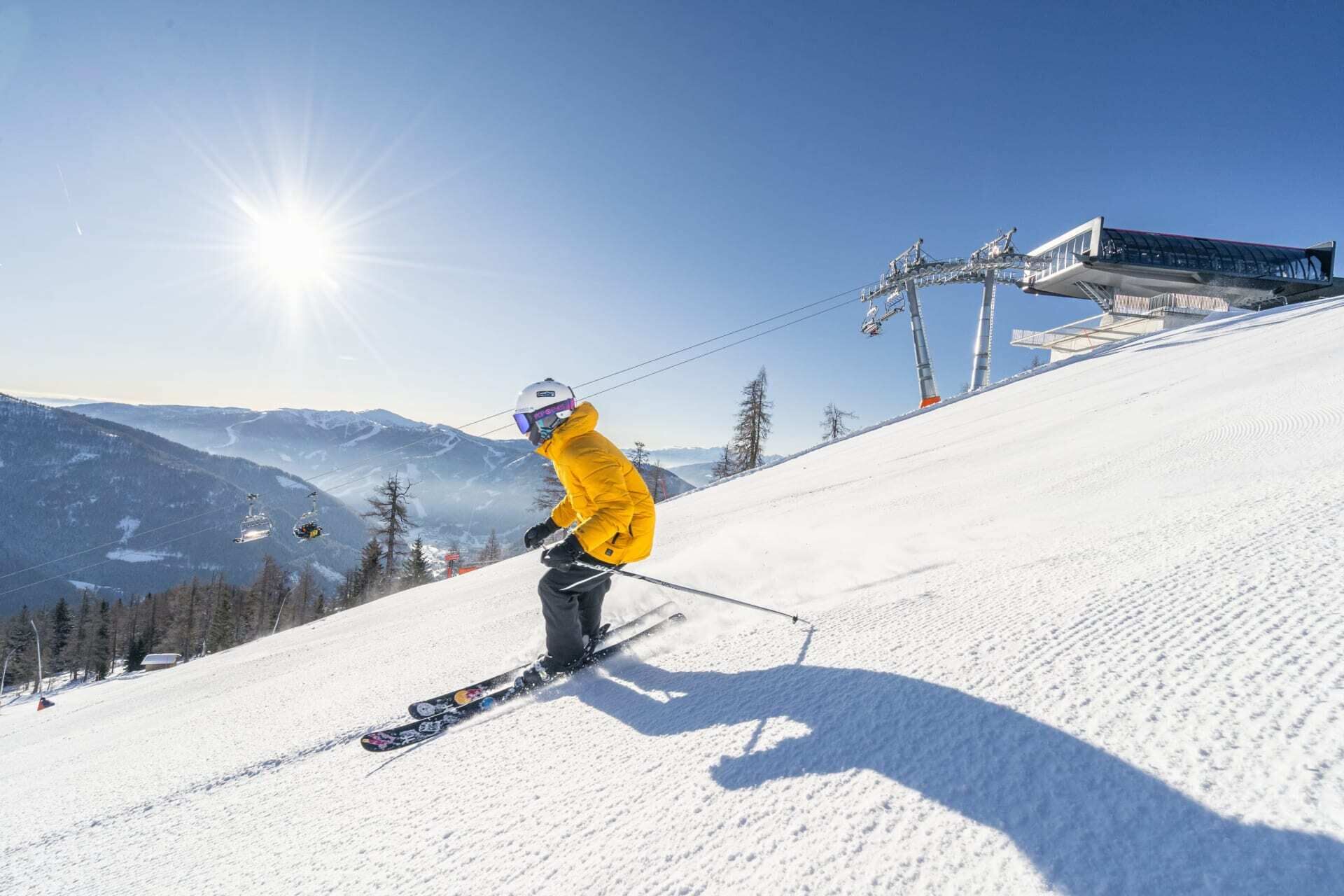 Skifahrer in gelber Jacke genießt sonnige Abfahrt auf schneebedecktem Hang unter blauem Himmel.