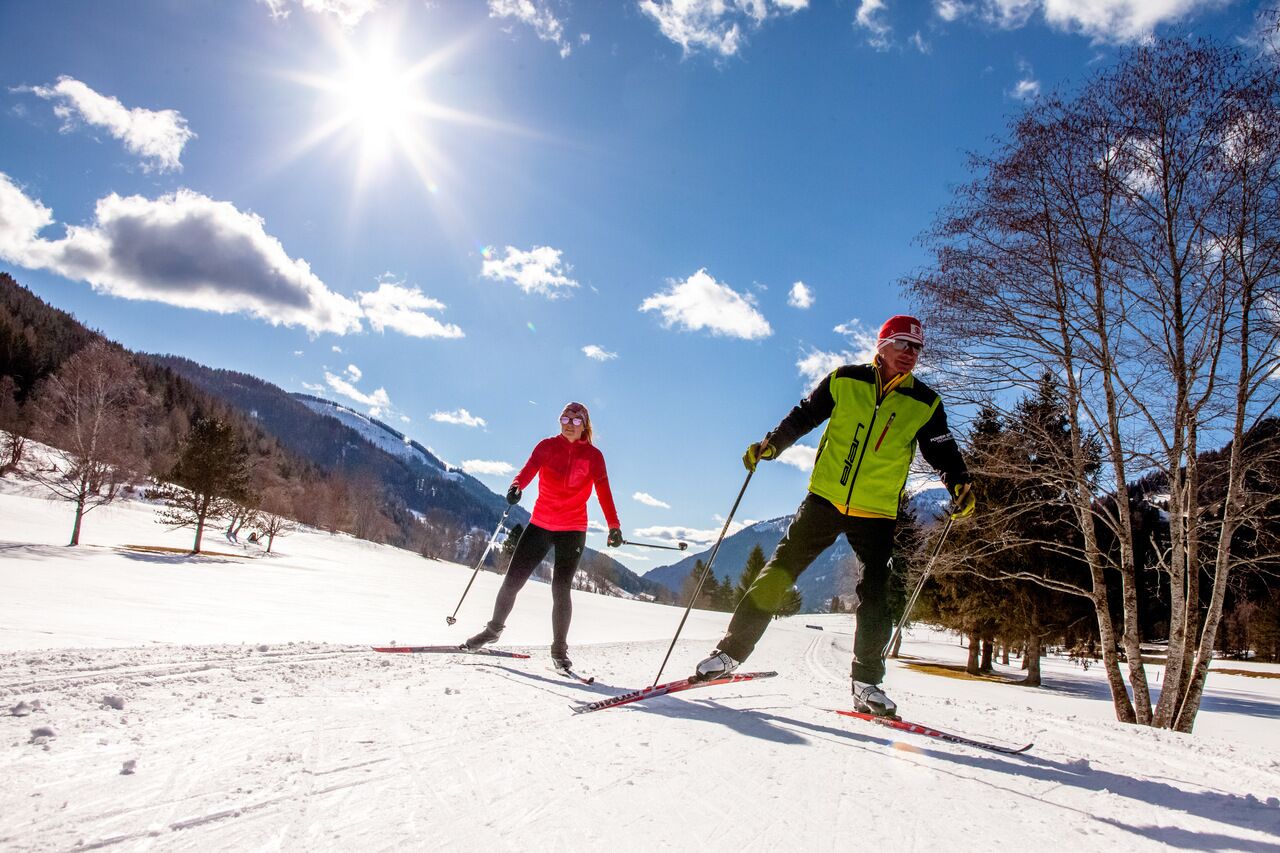 Two people cross-country skiing under a bright sky in a snowy mountain landscape.