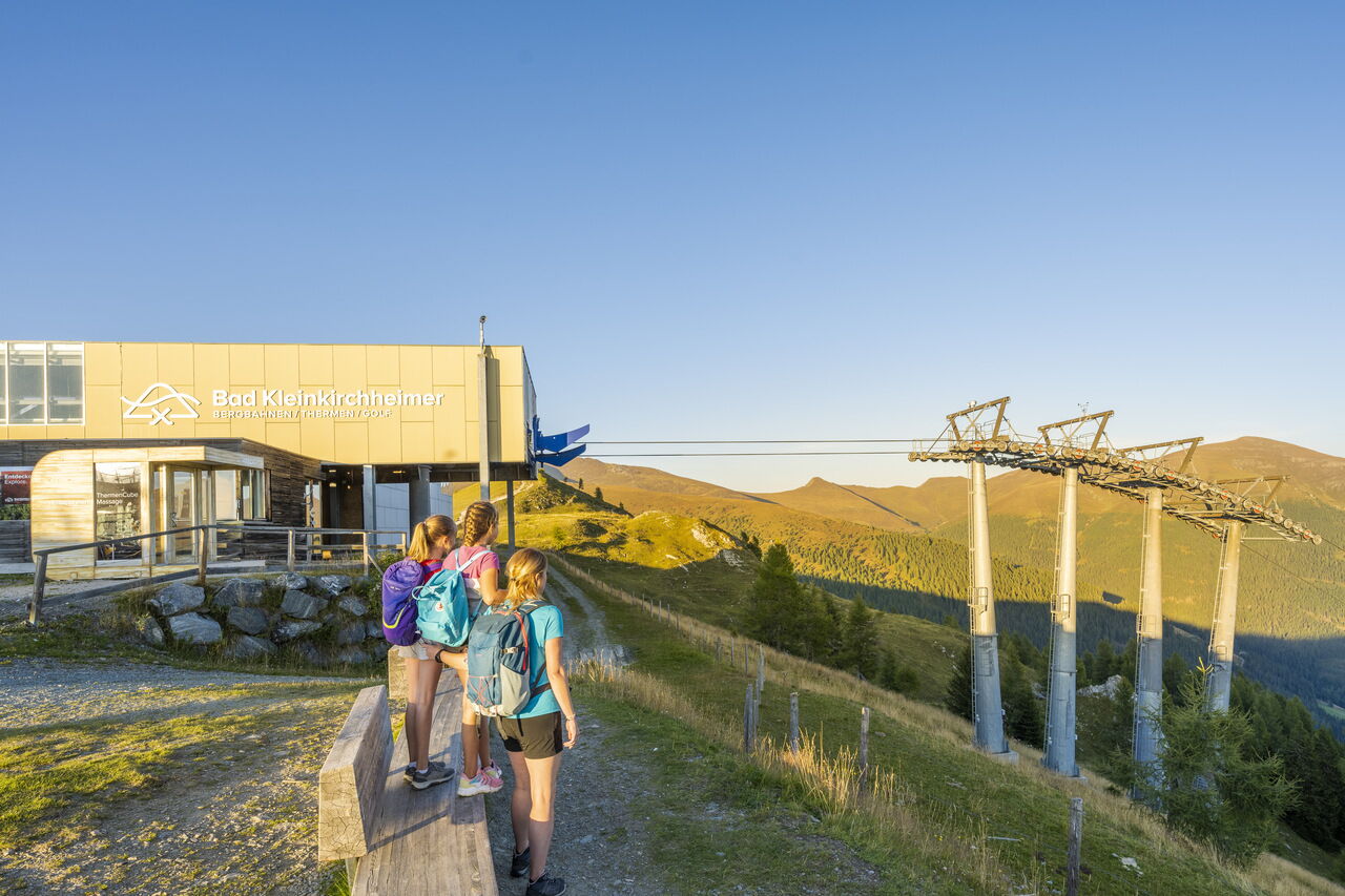 Drei Kinder mit Rucksäcken genießen die Aussicht bei sonnigem Wetter vor Bergstation.