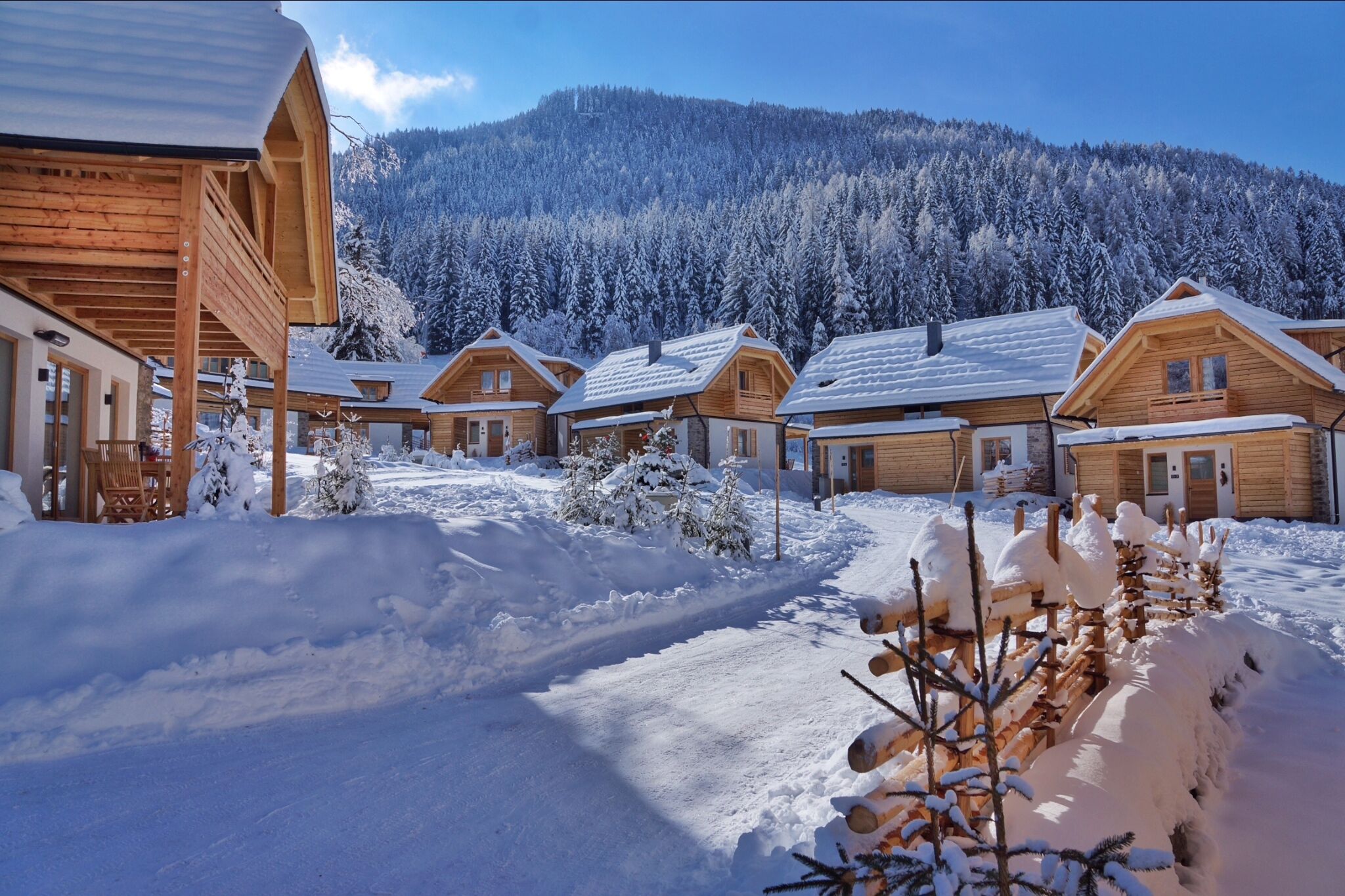A path that leads to several wooden huts in the snow next to a forest.