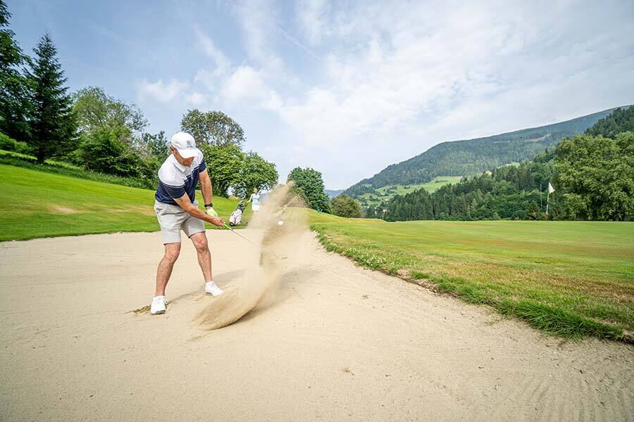 Golfer hits ball out of sand bunker on manicured green in sunny weather.