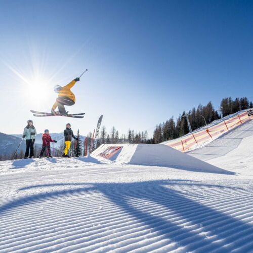 Skifahrer springt über Hügel, Familie schaut zu, blauer Himmel im Hintergrund.