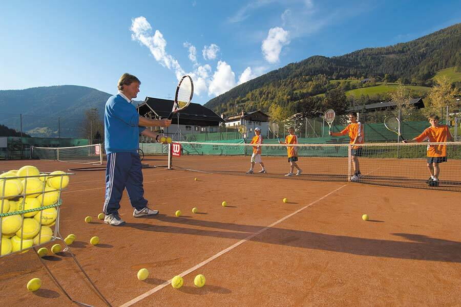 Lezioni di tennis su un campo in terra rossa, circondato da montagne pittoresche e cielo azzurro.